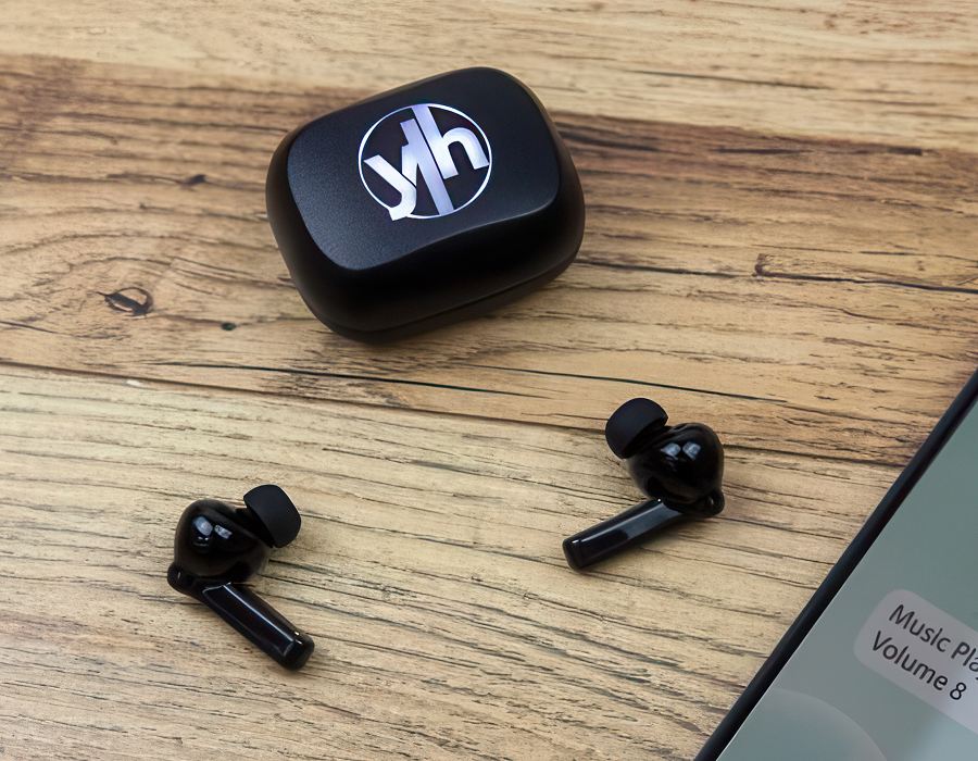 Black earbuds on a wooden table with illuminated case and part of a mobile phone screen showing music volume